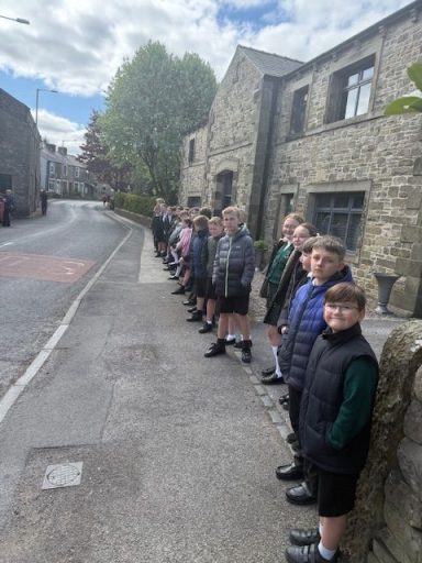 Children lined up on a rural road in front of stone buildings under a cloudy sky.