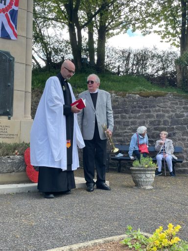 Clergy member in robes speaks to a man in sunglasses near a memorial, with seated people nearby.