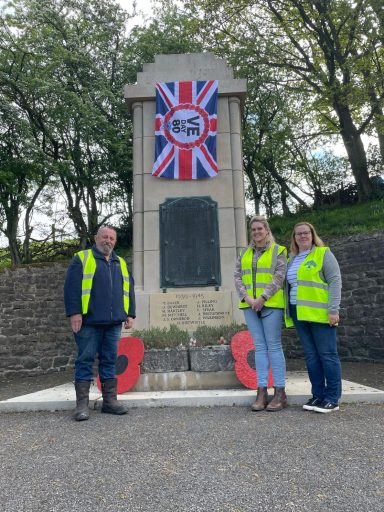 Three people in high-visibility jackets stand by a war memorial adorned with a Union Jack.