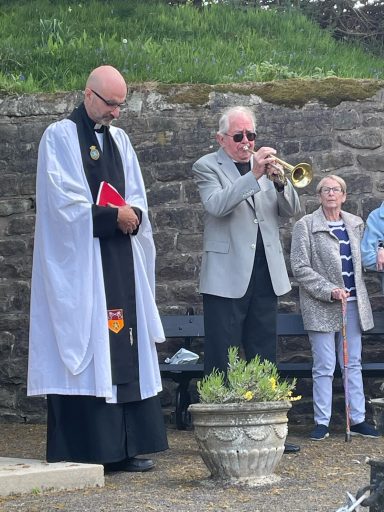 A priest stands with a Bible while a man plays trumpet; an elderly woman observes nearby.